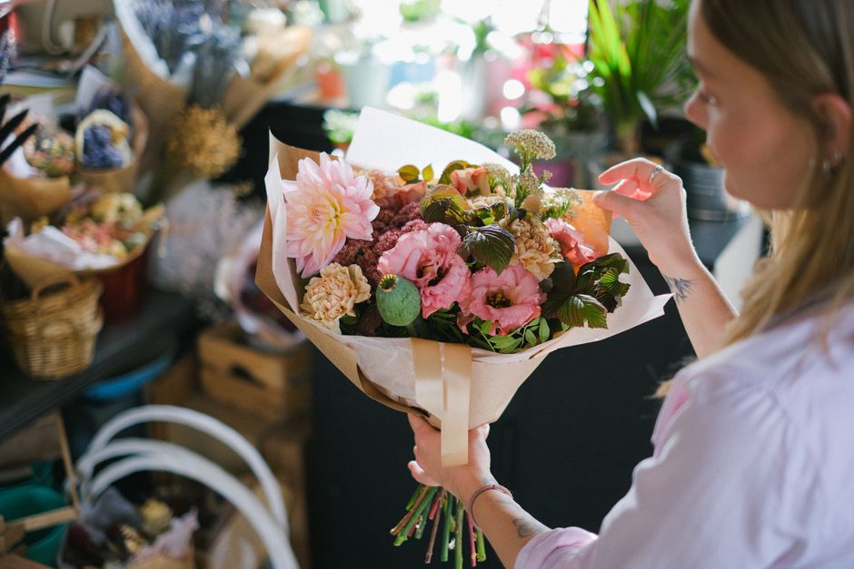 A woman with long blonde hair and light skin is holding a large bouquet of fresh flowers. The bouquet includes soft pink dahlias, delicate pink roses, light purple lisianthus, and green foliage wrappe