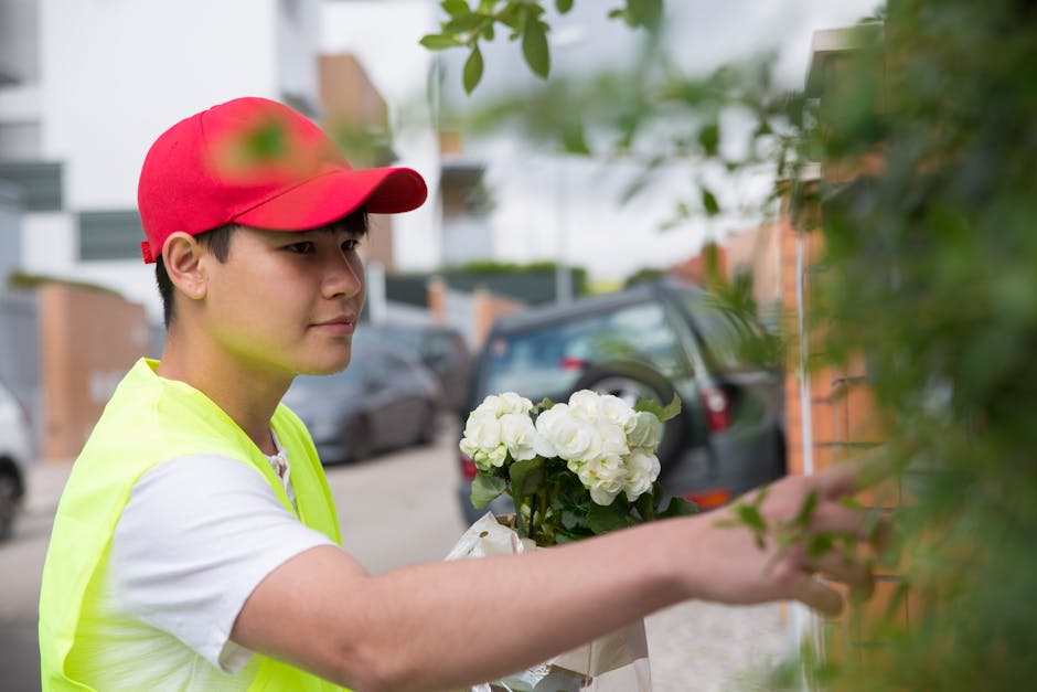 A young male florist wearing a red cap and a high-visibility yellow vest is arranging or delivering a bouquet of fresh white flowers, possibly hydrangeas or roses, wrapped in light-colored paper with