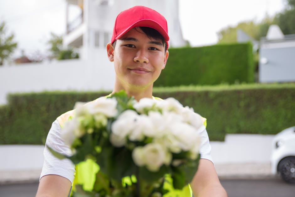 A delivery person wearing a red cap and high-visibility vest reaching out with a bouquet of fresh white flowers, including roses and other blooms, wrapped in clear cellophane and tied with a light-col
