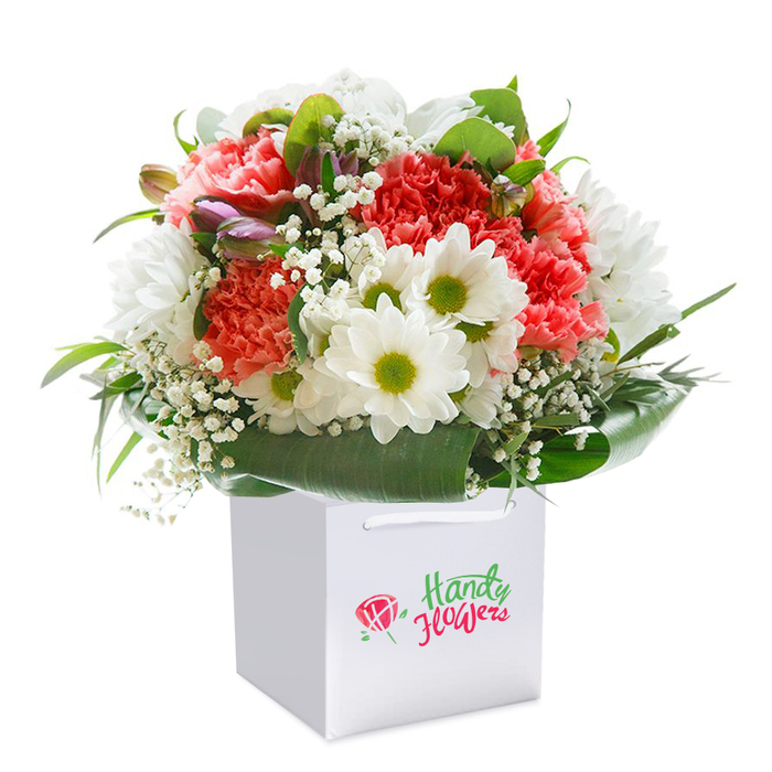 A vibrant hand-tied bouquet is pictured upright in a flawless white gift bag, exuding a fresh, celebratory spirit that feels at home in Malden Rushett's tranquil surrounds. The arrangement is rich with ruffled carnations in coral, peach, and red-each bloom bursting with colour and texture, their velvety petals inviting both the eye and imagination. Daisies in bright white with radiant yellow-green centres lend natural cheer, their dainty forms reminiscent of wildflowers picked along Rushett Lane on a sunny day. Wispy baby's breath is gently threaded throughout, adding a light, cloudlike finish that softly blurs the boundaries between each main flower. Along the left side, a handful of purple-tinged buds contribute depth and subtle intrigue, symbolising the new beginnings that abound in springtime at Ashtead Common. A plentiful sweep of glossy, deep green foliages envelopes the base, elegantly supporting the arrangement while suggesting the untamed greenery that edges local gardens. Secured by sturdy white ribbon handles, the square white bag is elegant and practical for convenient, same-day floral delivery across Malden Rushett and beyond. Set against an immaculate white background, the image highlights not just the flowers' beauty and freshness, but also the thoughtful care of the local artisans who assemble each bouquet. This artistic floral presentation is perfect for birthdays, anniversaries, homecomings-or simply to send warm wishes throughout the Malden Rushett community.